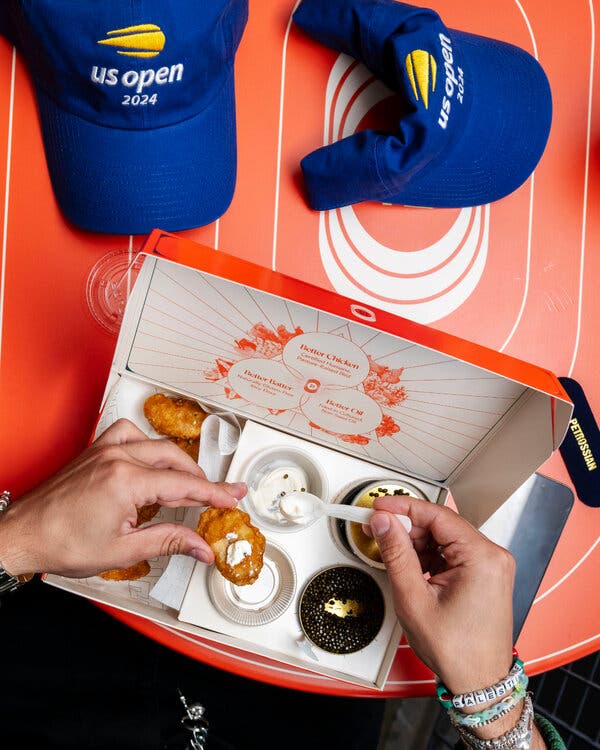 Two hands are seen spooning crème fraîche onto a chicken nugget which is in an orange box. The box is sitting on an orange table with two blue U.S. Open hats.