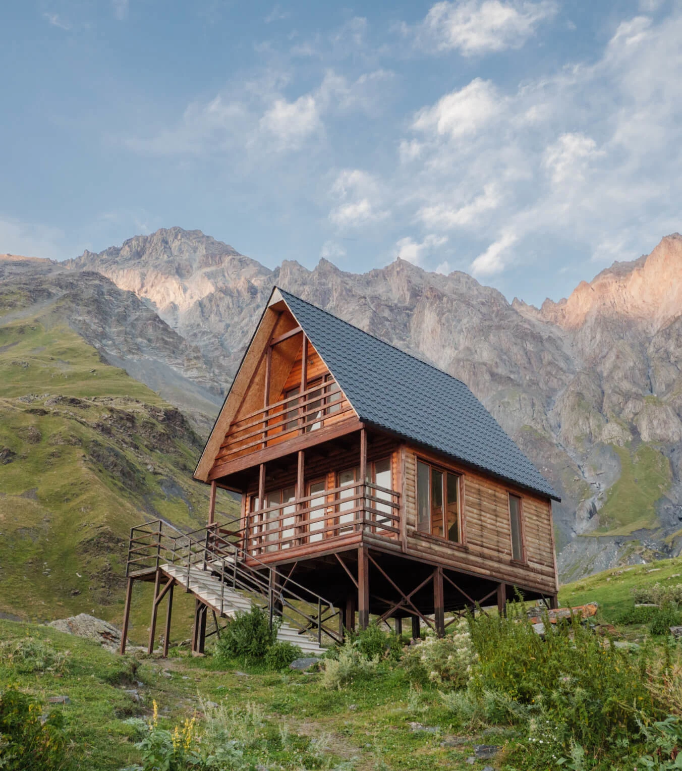 A rustic wooden cabin sits on stilts. Behind, there’s a scenic mountain backdrop with grassy hills and a blue sky.